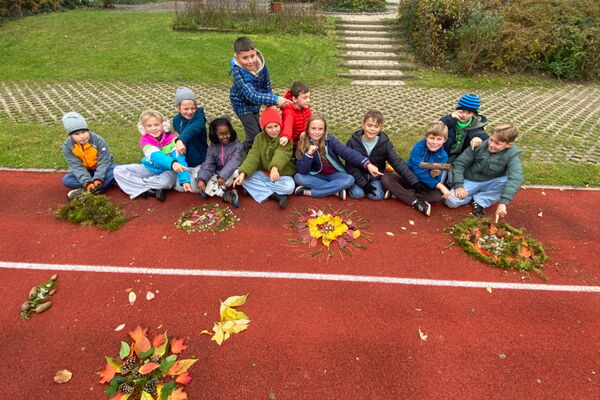 Schulkinder sitzen mit bunten Kränzen aus Naturmaterialien auf dem Hartplatz